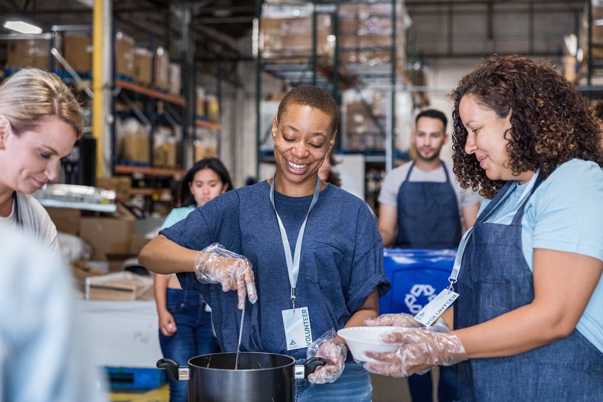 Volunteers wearing aprons and gloves prepare and serve food together inside a warehouse-style community space.