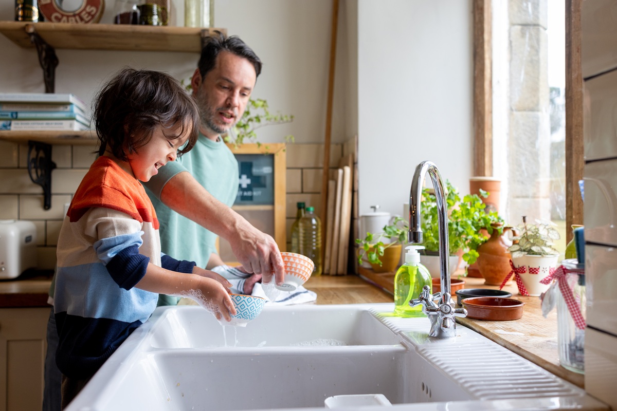 An adult and a young child wash dishes together at a bright kitchen sink with sunlight and plants nearby.