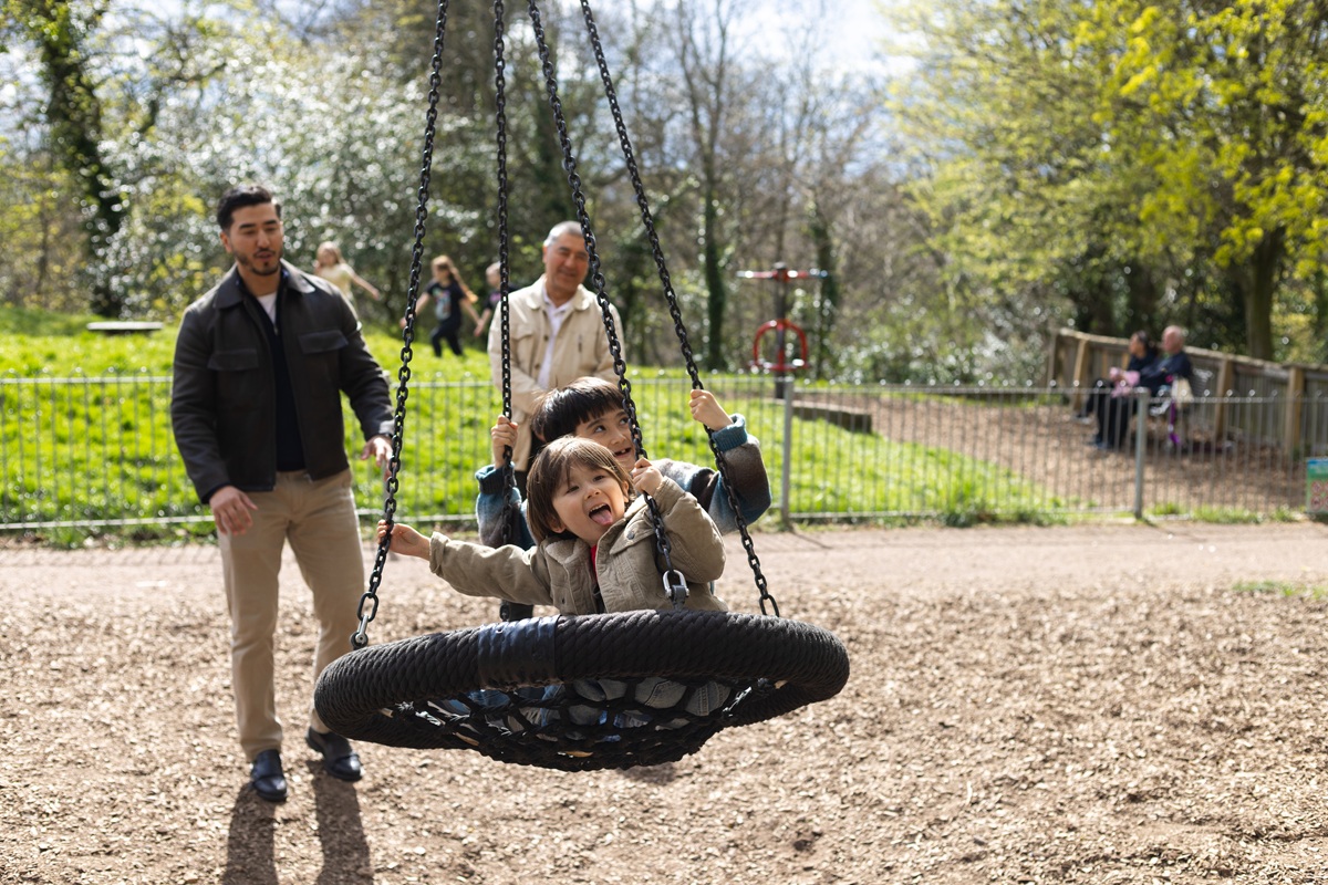 Adults stand near a playground swing as a child rides a circular net swing in a sunny park with trees and a fenced play area in the background.
