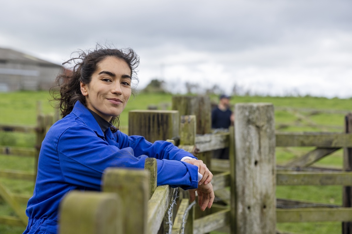 A confident woman in a blue jacket leans on a wooden fence in a green, open field.