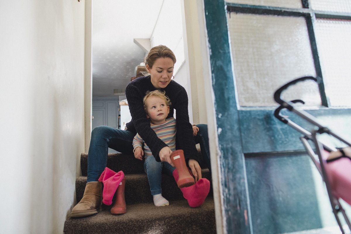 A parent helping a young child put on bright pink rain boots while sitting on a staircase at home.