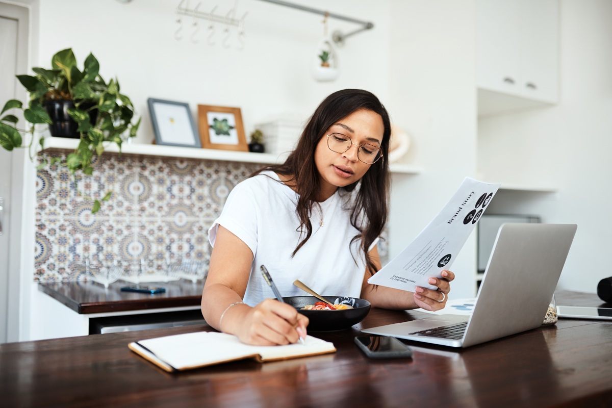 Person working at a kitchen table with a laptop, documents, and a notebook.