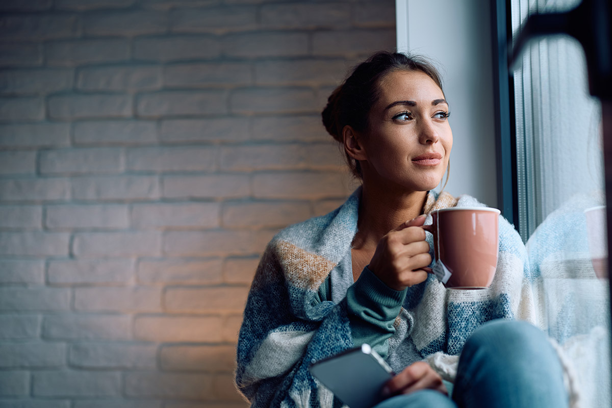 Individual sitting by a window holding a coffee mug and mobile phone, reflecting on financial well being in a home setting.