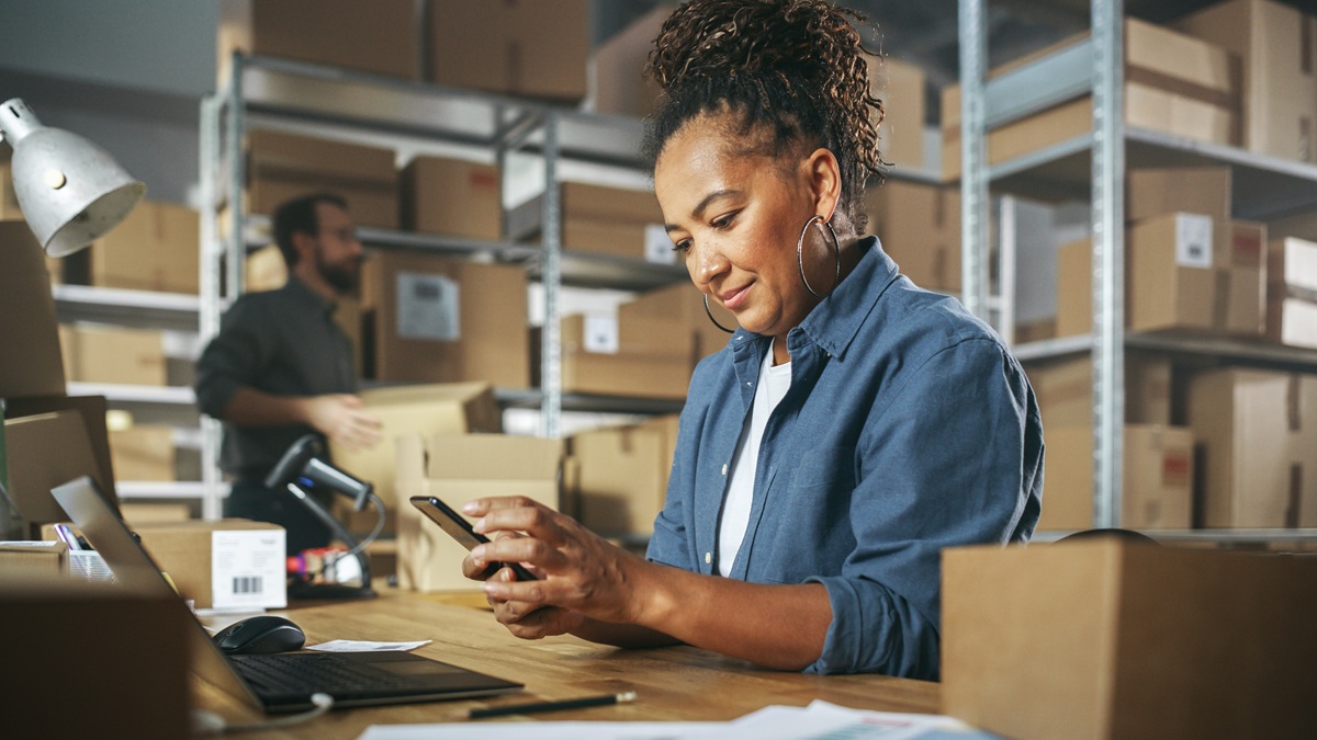 A small business owner checks a smartphone while working at a desk in a warehouse, with shelves of packaged boxes and a coworker handling inventory in the background.
