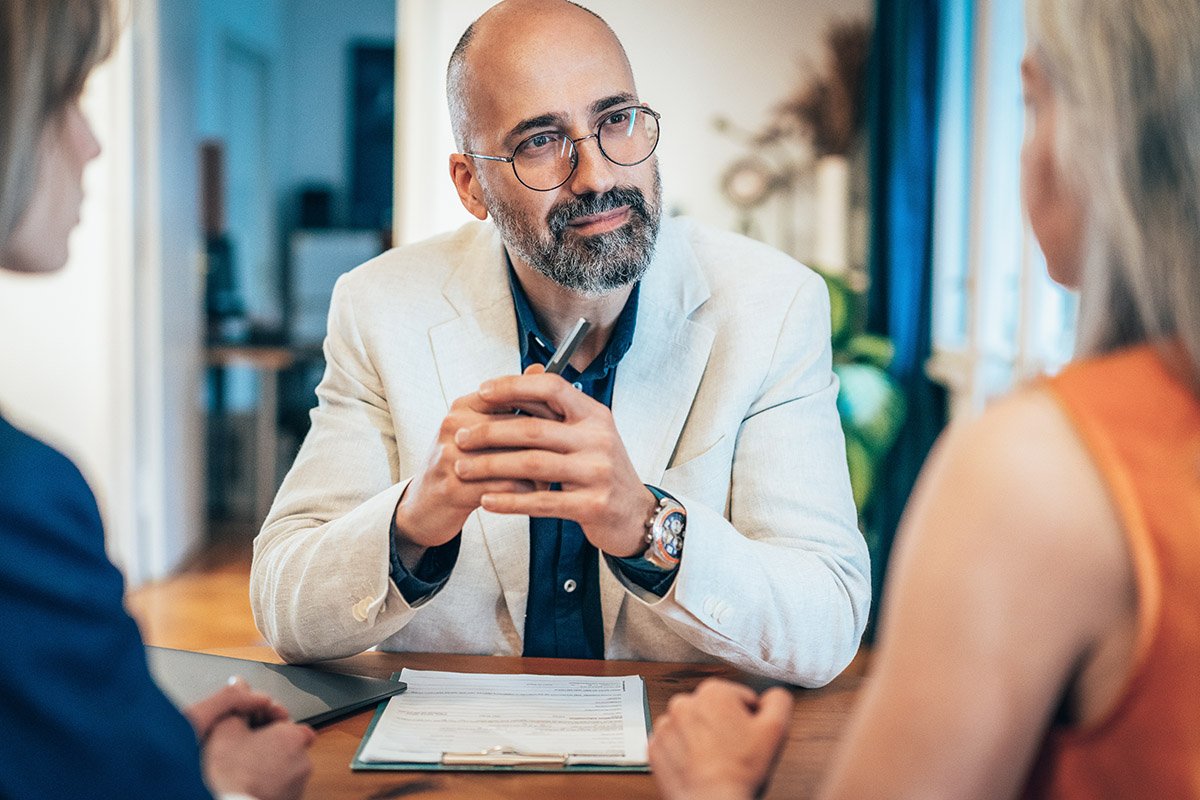 A professional advisor sits at a table holding a pen while meeting with two clients, reviewing documents during a consultation in a modern office setting.