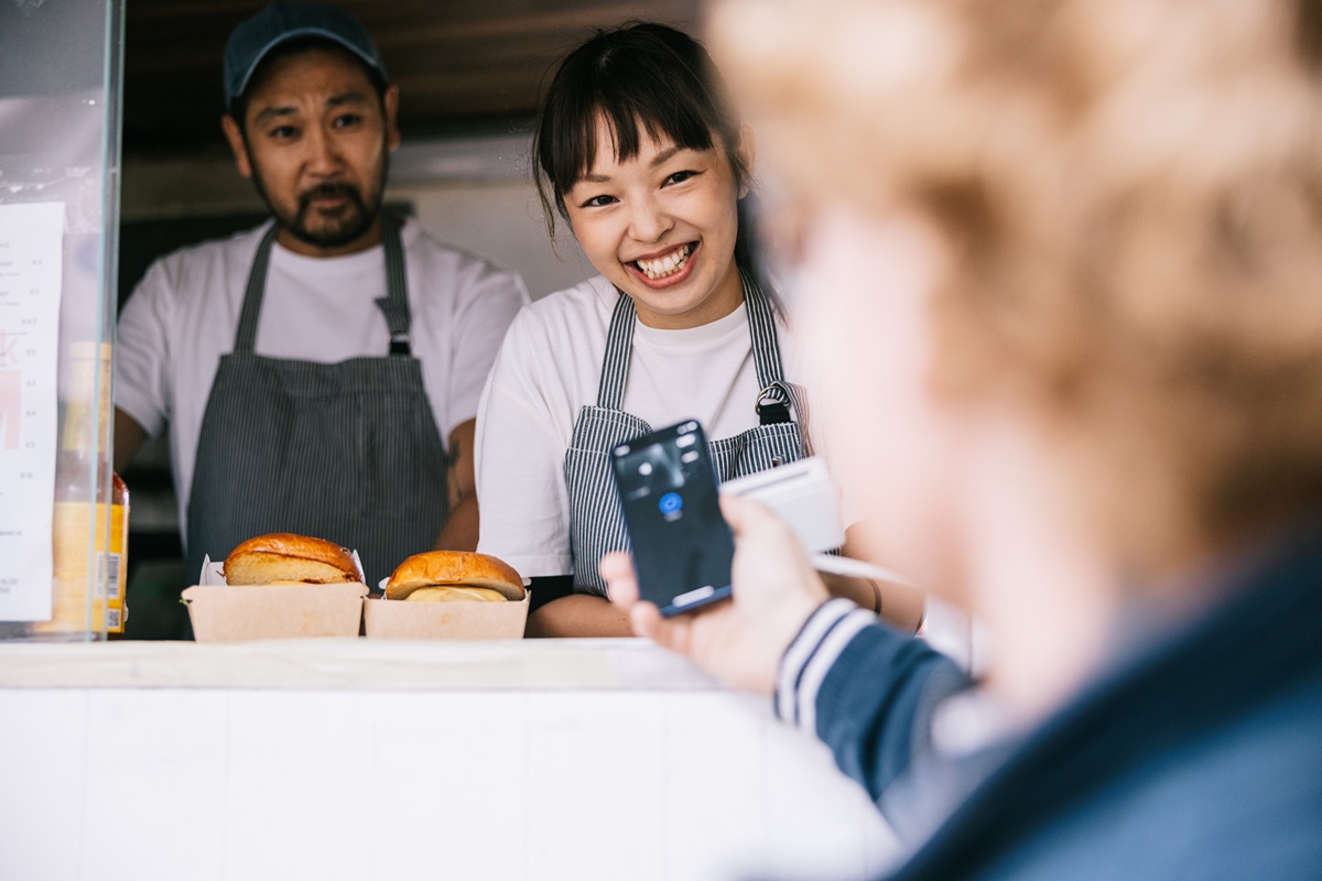 A customer uses a smartphone for contactless payment at a food stand, with two staff members in aprons behind the counter and sandwiches displayed in front of them.