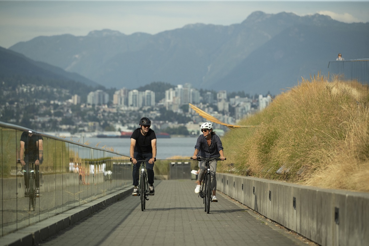 Two people ride bicycles along a waterfront pathway with Vancouver’s skyline and mountains in the background on a sunny day.