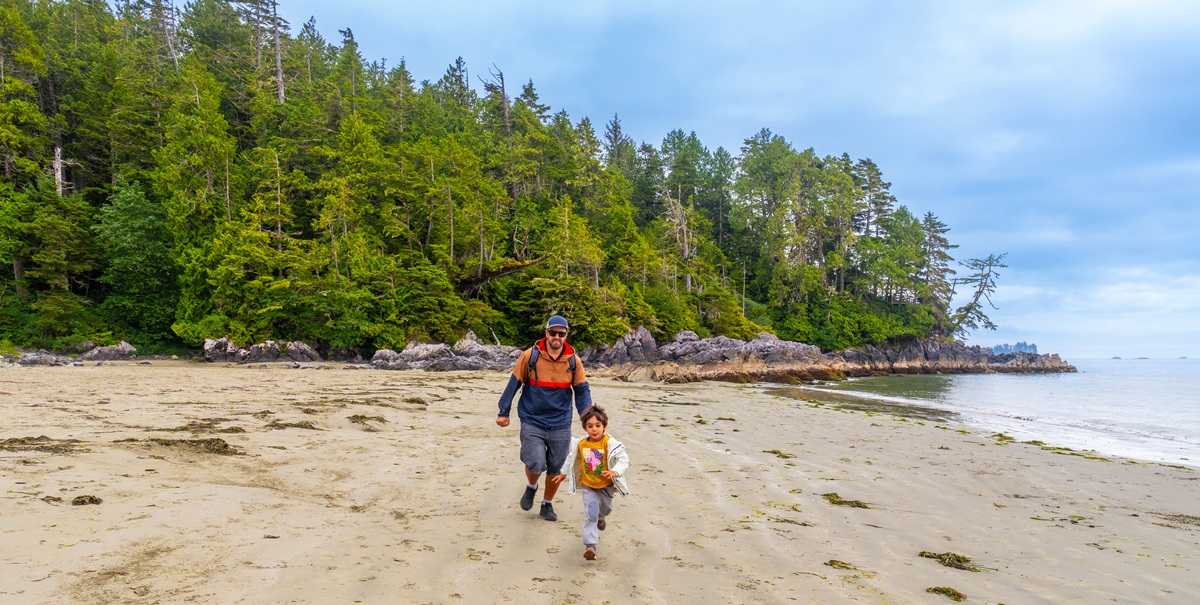 Adult and child walking along a sandy beach with forested shoreline and calm water nearby.