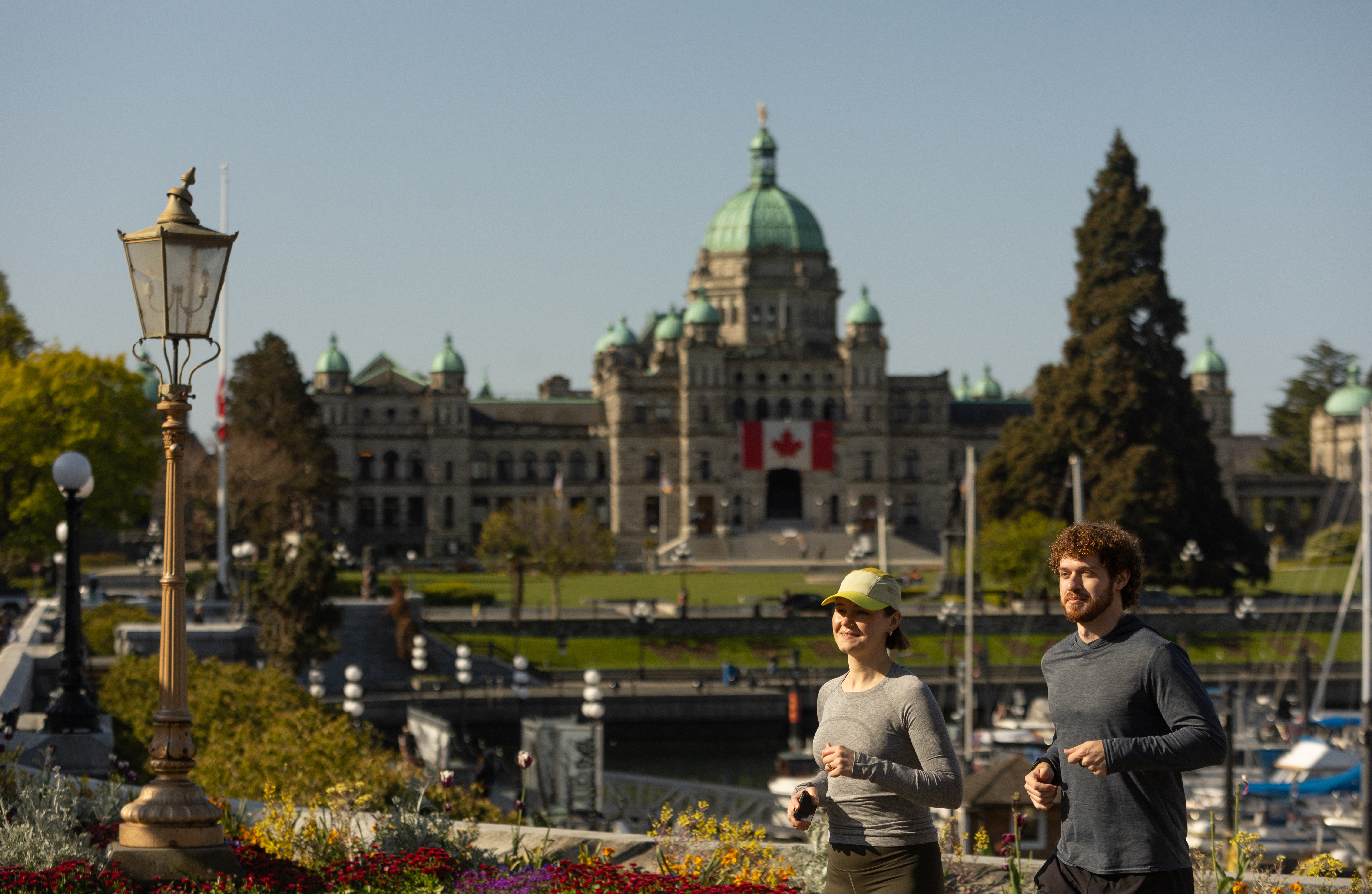 Two people jog along a waterfront path in front of the BC parliament building with a Canadian flag, with gardens, boats, and trees visible under a clear blue sky.