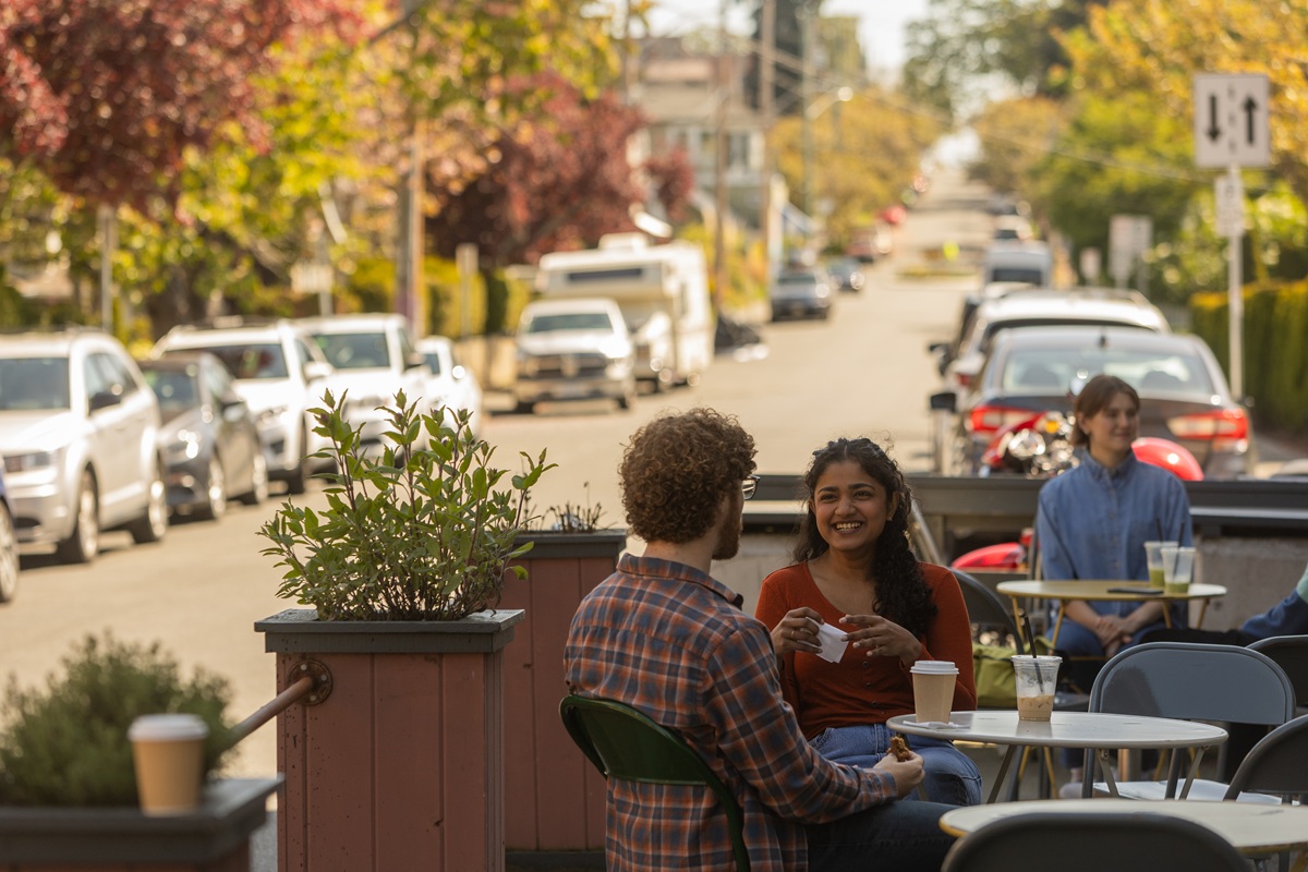 People enjoying coffee at an outdoor café on a sunny street lined with parked cars and trees.