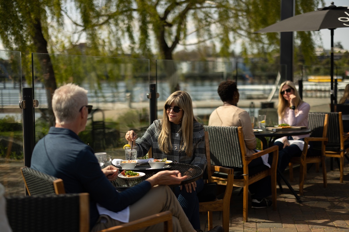 People enjoying a meal together on an outdoor patio beside the water on a sunny day.