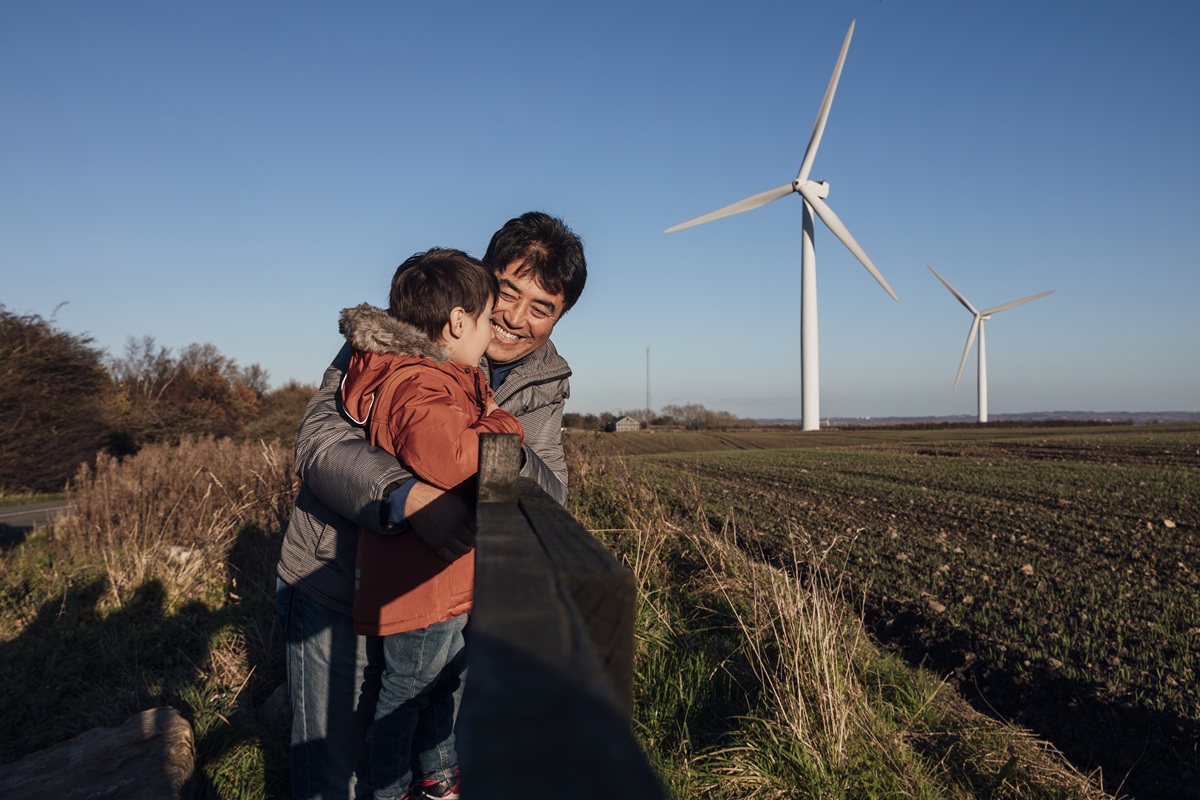 Two people stand by a wooden fence overlooking open farmland with wind turbines under a clear blue sky.
