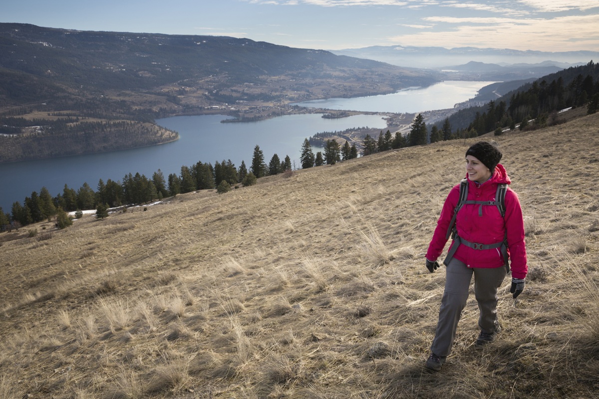 Hiker in a red jacket walking across a grassy hillside overlooking a winding lake and mountain landscape.