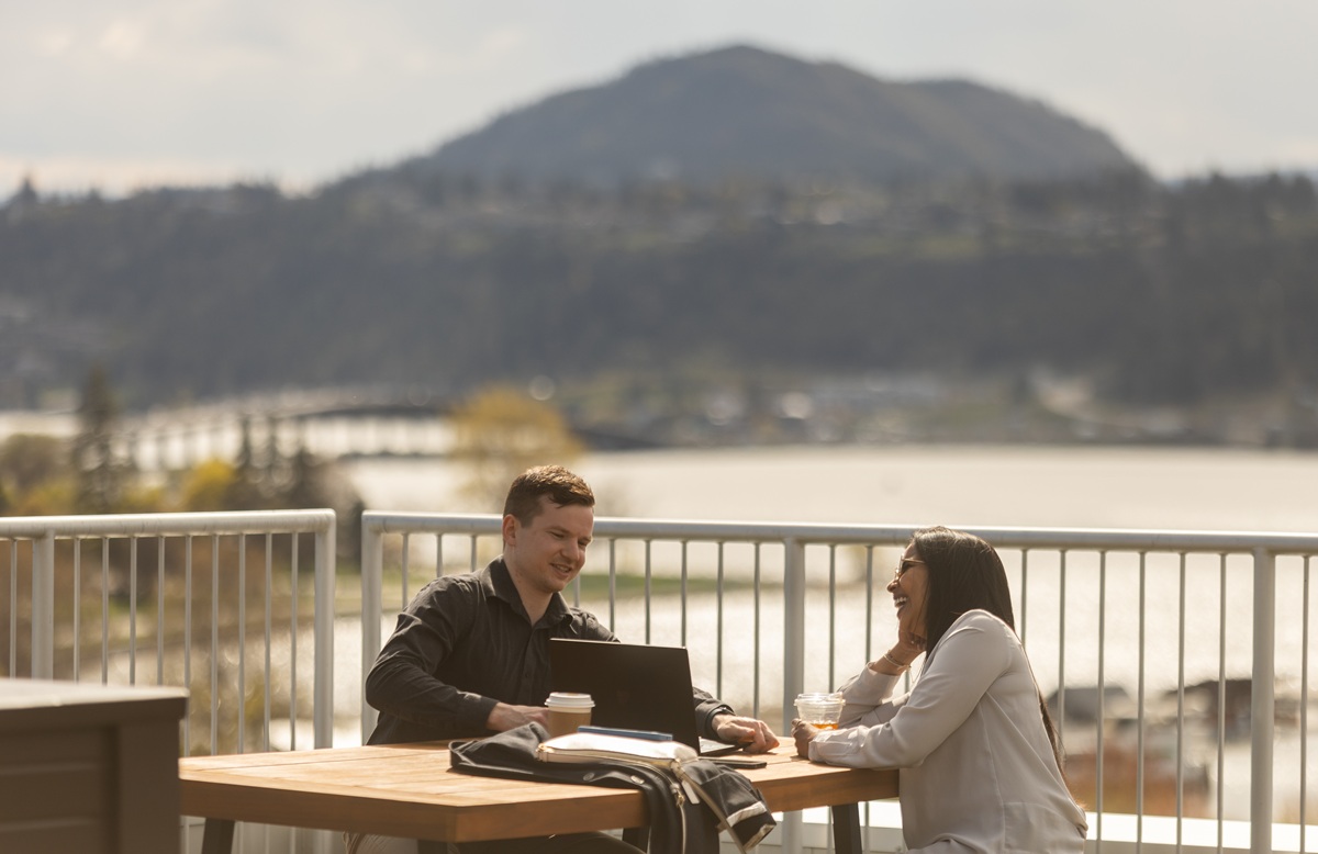 Two people sit at an outdoor table by the water, talking over coffee and a laptop with mountains and a lake in the background.