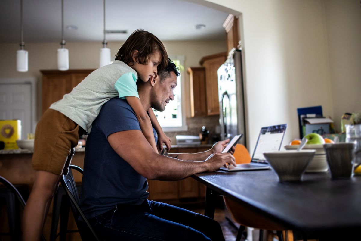 An adult sits at a kitchen table using a tablet while a young child hugs them from behind, with a laptop and fruit bowl on the table.