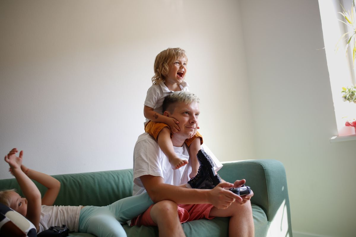 An adult sits on a green sofa holding a game controller while two young children play nearby, one perched on their shoulders and another lying back on the couch in a bright, sunlit living room.
