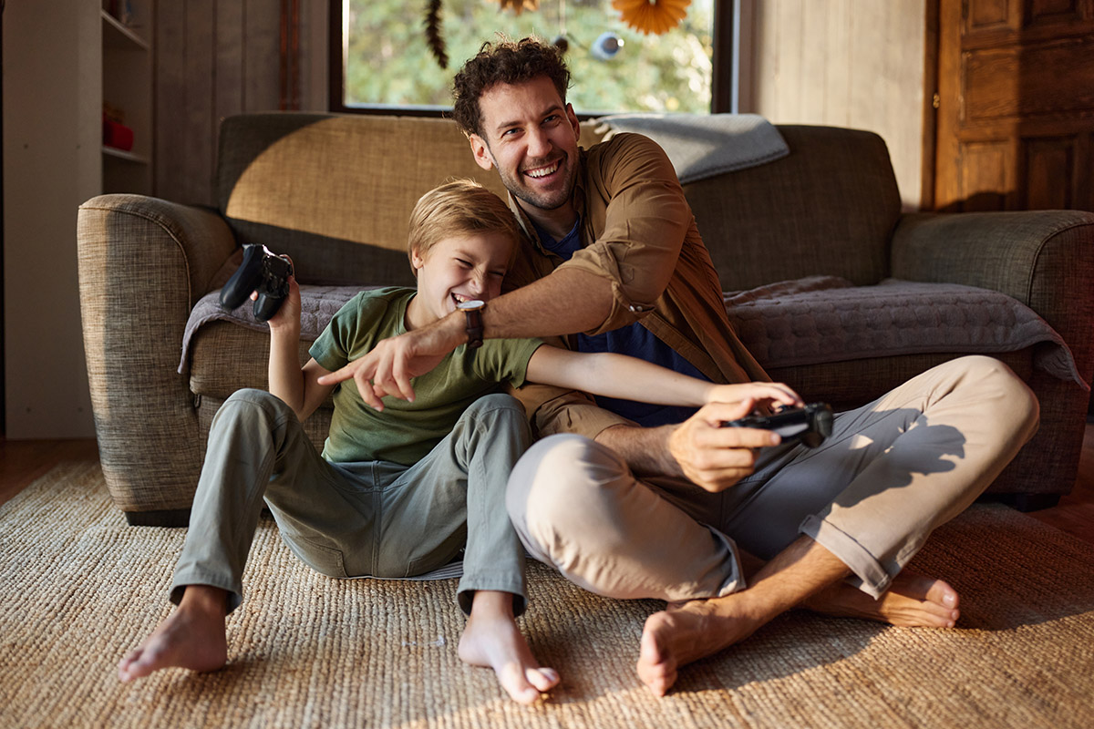 An adult and a child sit on the floor against a sofa, holding video game controllers and playing together in a warmly lit living room.