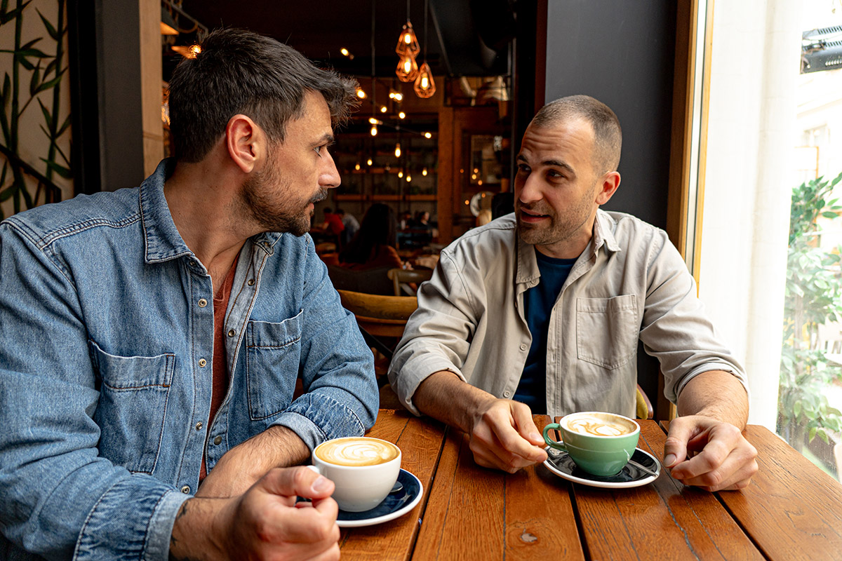 Two people sit at a wooden table in a café, talking over cups of coffee near a window, with warm lighting and a relaxed atmosphere.
