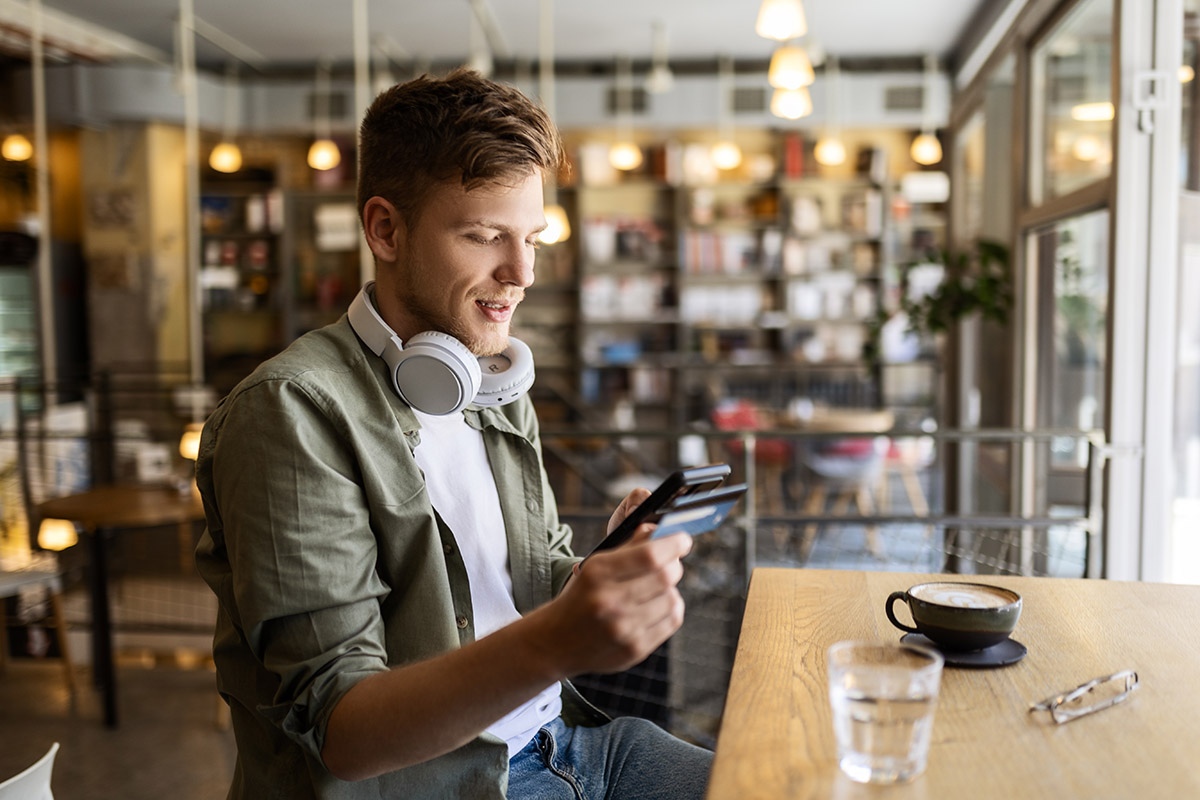A person sits at a wooden table in a cozy café, wearing a light jacket and headphones around their neck while looking at a smartphone.
