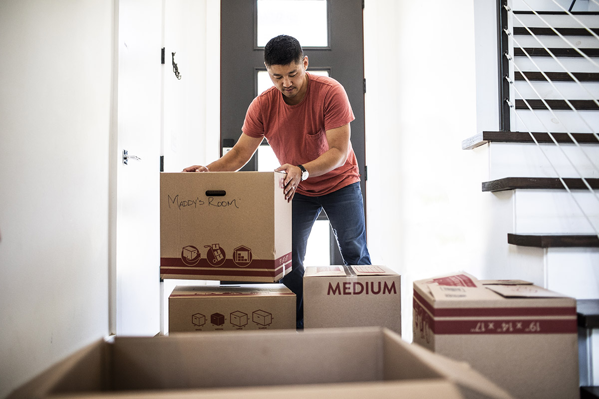 A person stands in a bright entryway stacking and organizing cardboard moving boxes near the front door, with a staircase visible to the side, suggesting a move or unpacking process.
