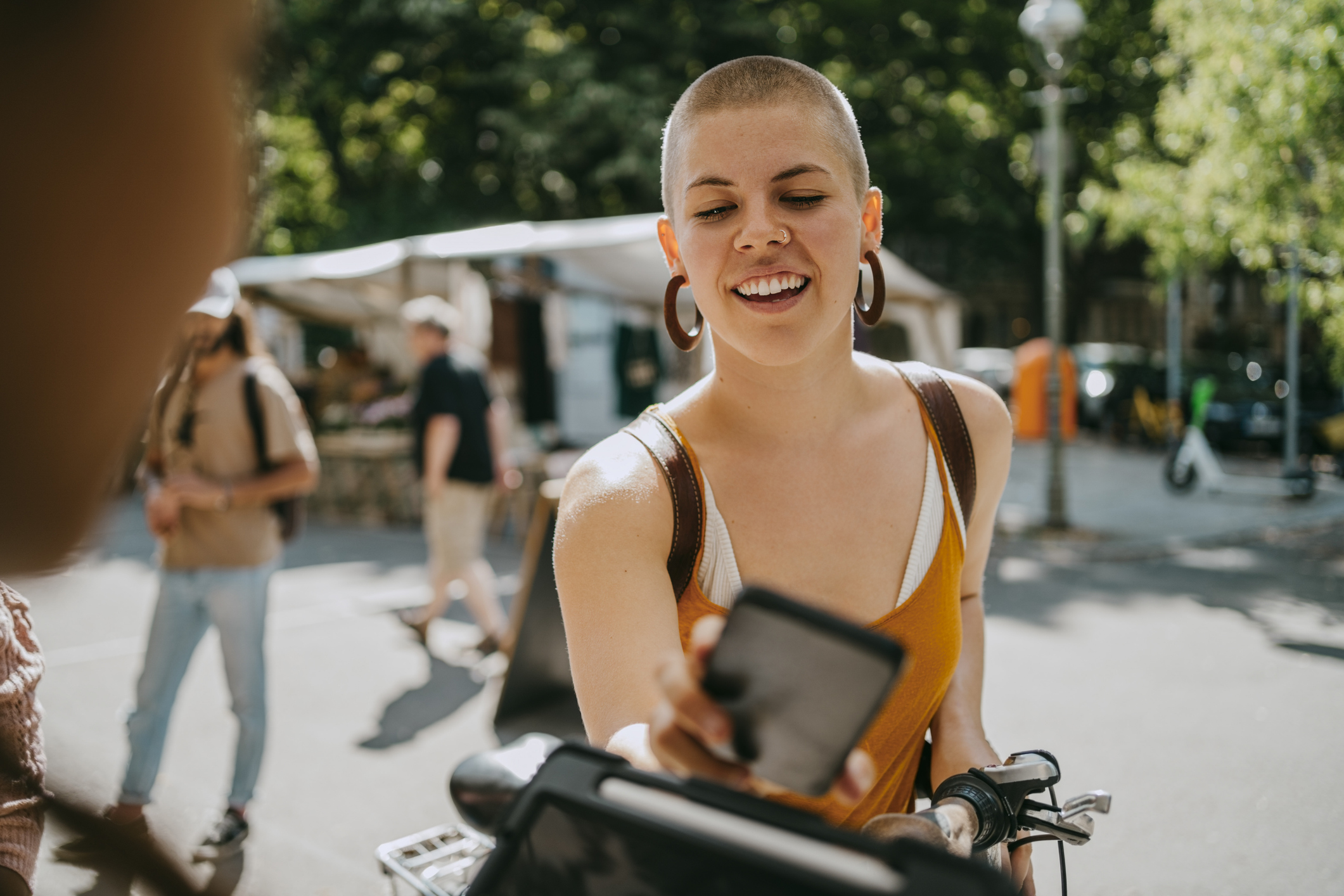 A person standing beside a bicycle extends a smartphone toward another device at an outdoor market or plaza, with people and stalls in the background.
