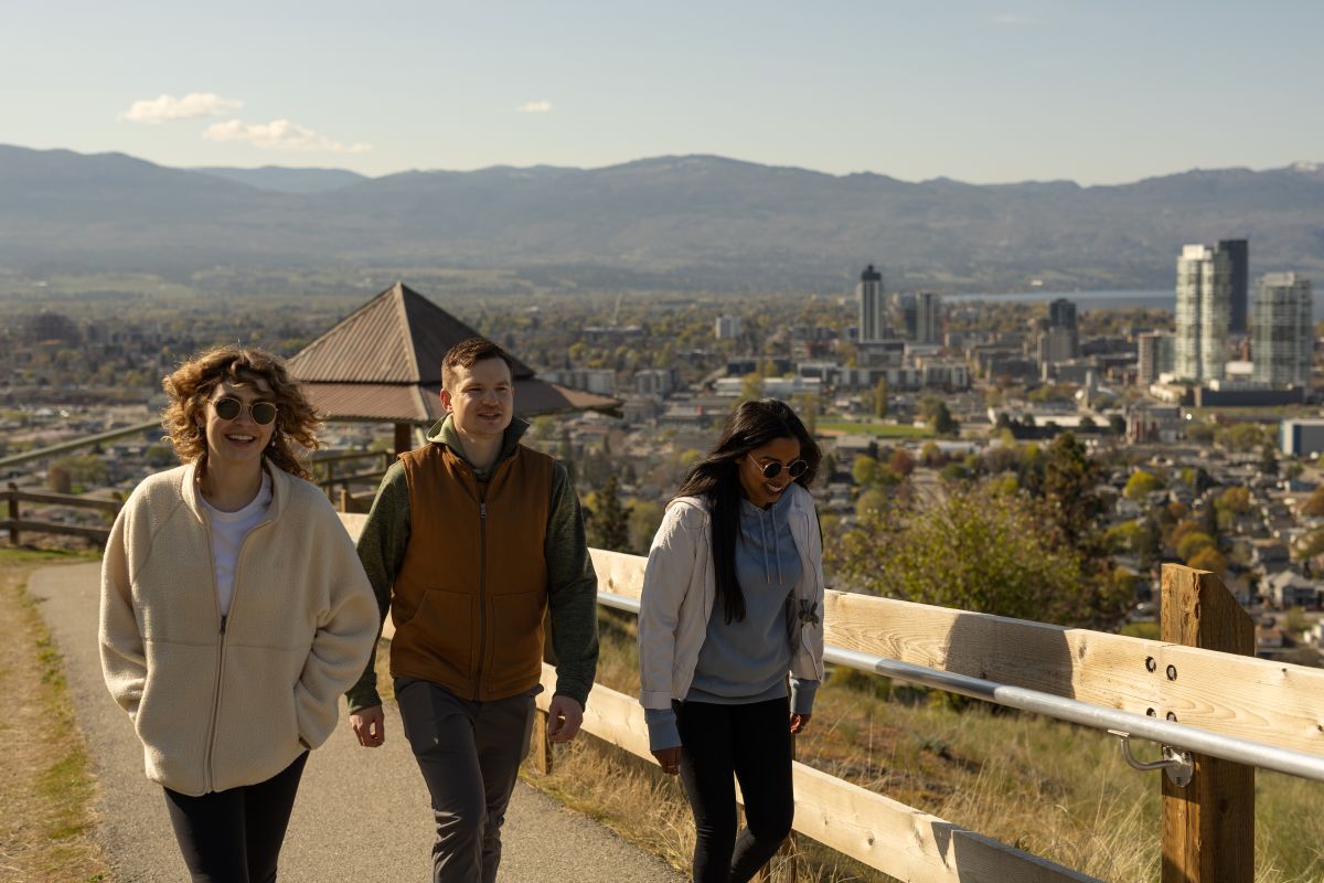 Three people walk along a paved hillside path beside a wooden railing, overlooking a sunlit city with high-rise buildings, tree-filled neighborhoods, and mountains in the distance under a clear sky.