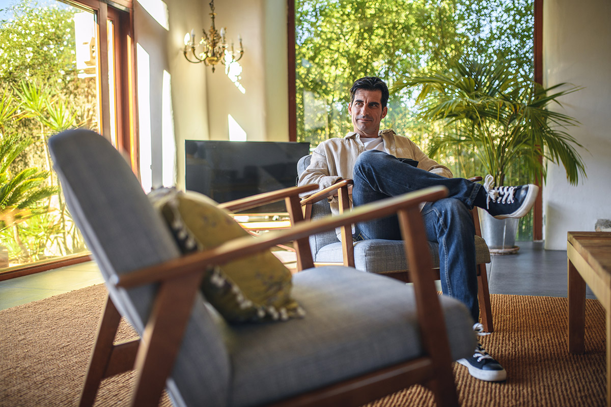 A person relaxes in a mid-century–style armchair inside a sunlit living room, with large windows, indoor plants, and warm wooden furniture creating a calm, airy atmosphere.